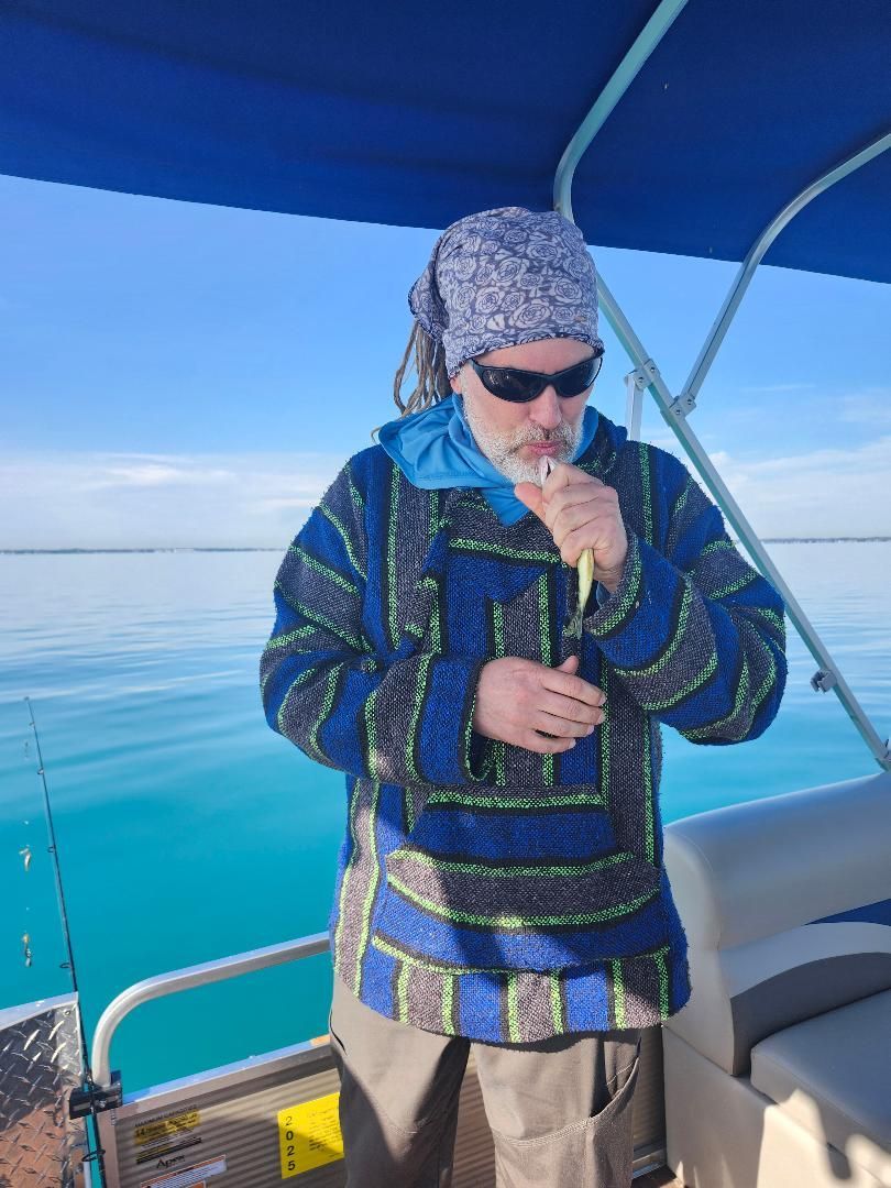 Man on a boat, wearing sunglasses and a patterned blue jacket, holding a small fish, with a blue sky background.