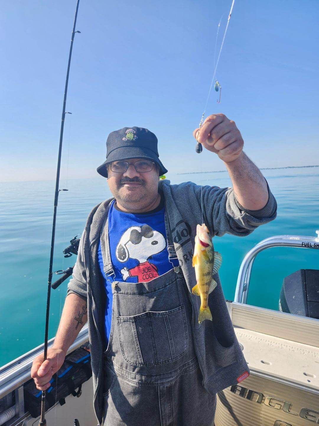 Man fishing on a boat, holding up a small yellow fish, blue water, sunny day.
