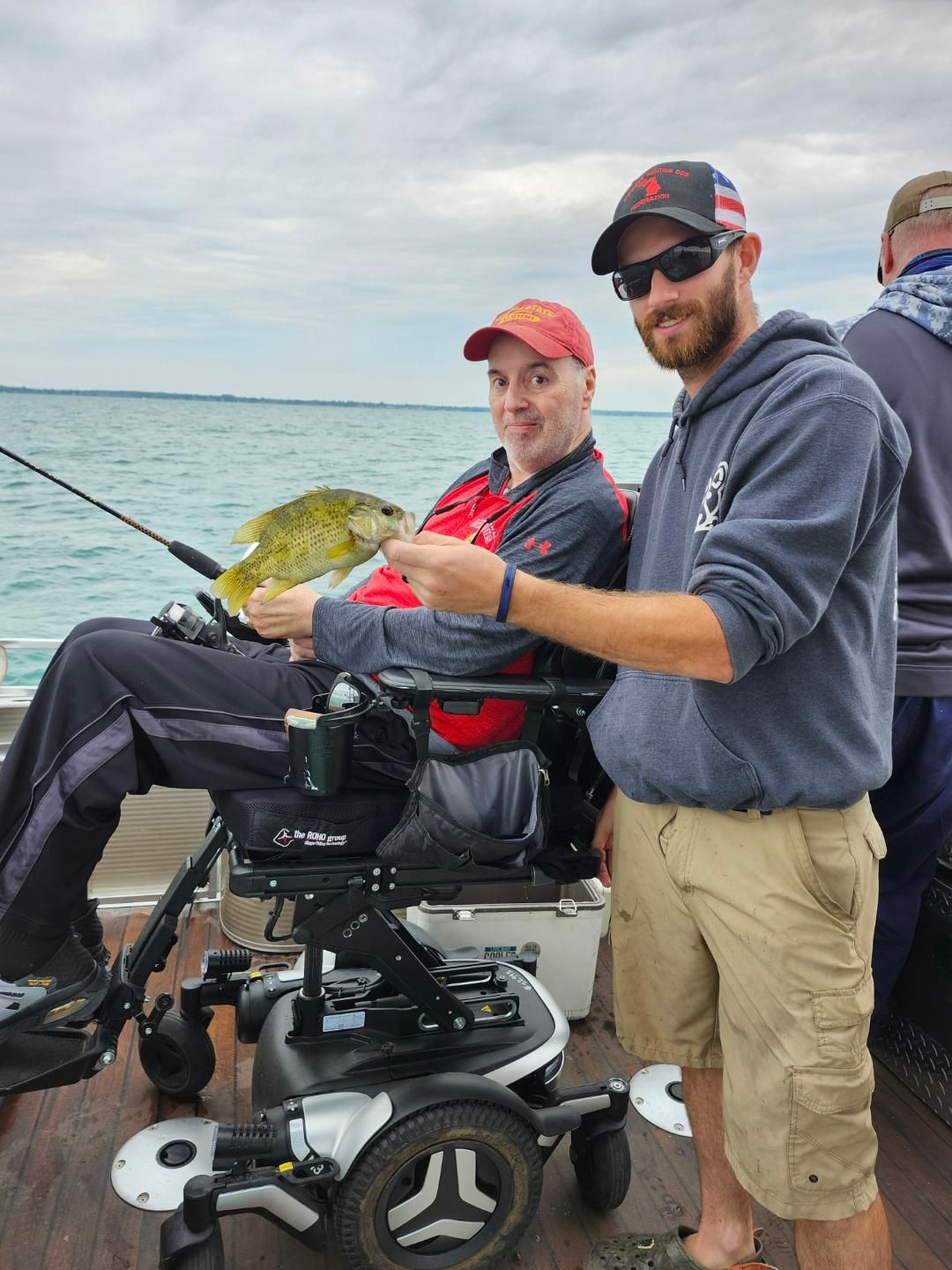 Man in wheelchair fishing on a boat, holding a fish, with a man standing beside him.