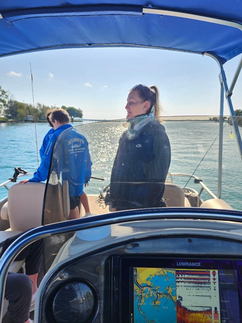 Woman on a boat, wearing a jacket, looking at the water. Two people fish in the background. Sunny day, open water.