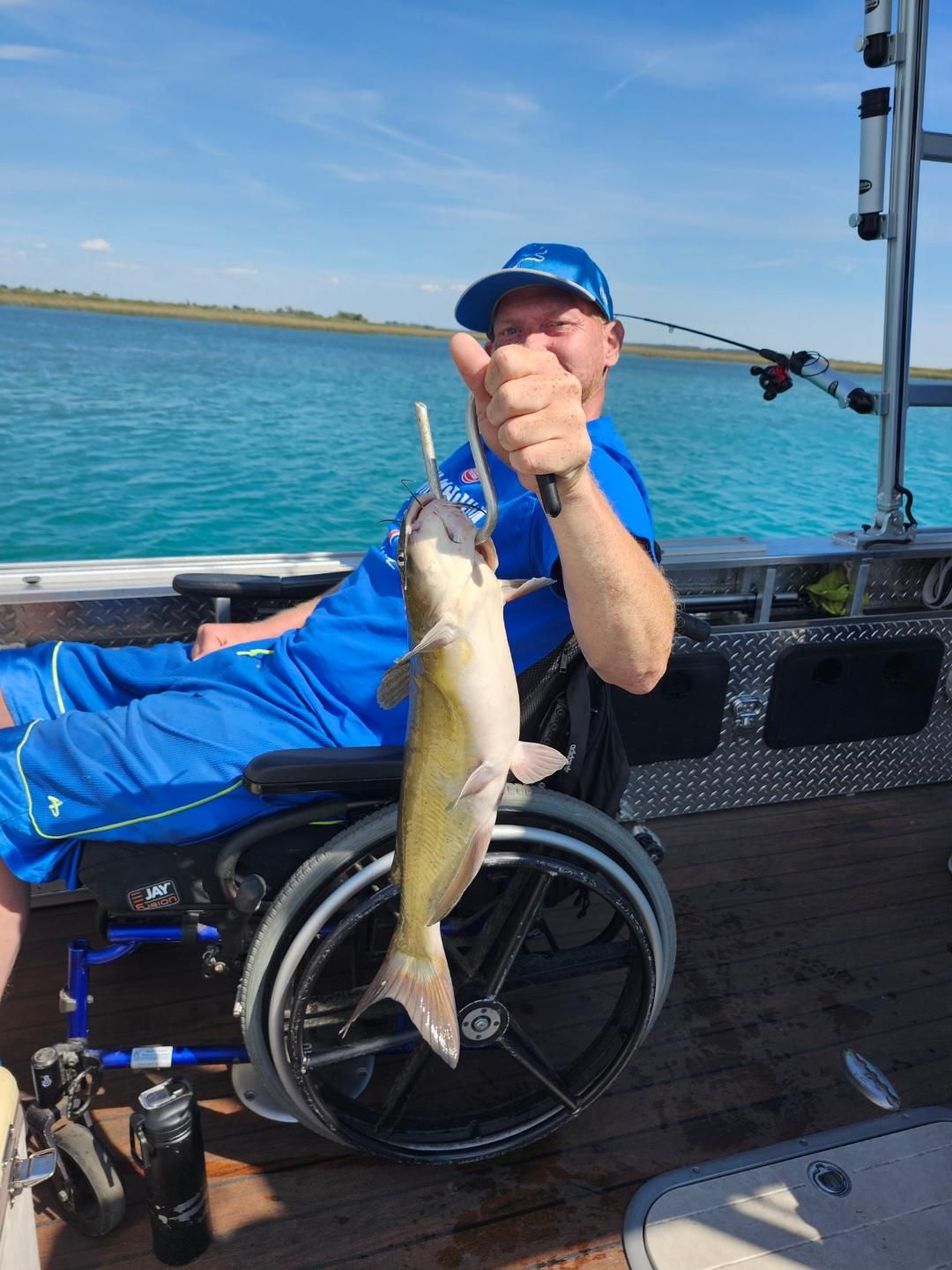 Man in wheelchair fishing, holding up a catfish. He gives a thumbs up on a boat with blue water in the background.