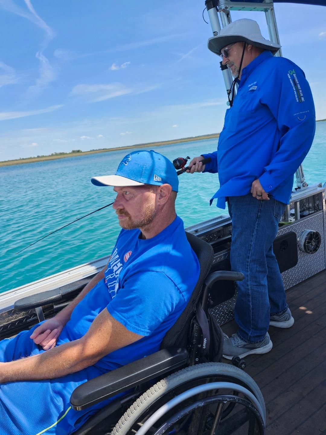 Man in wheelchair fishing on a boat with another person assisting; blue water and sky.