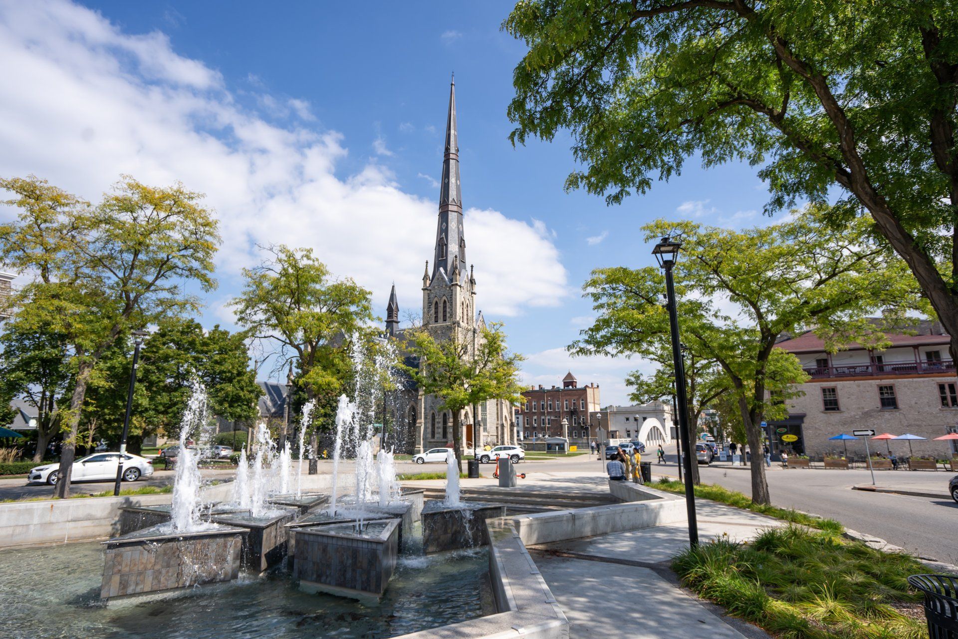 Fountain in front of a church with a tall spire on a sunny day. Trees and buildings line the street.