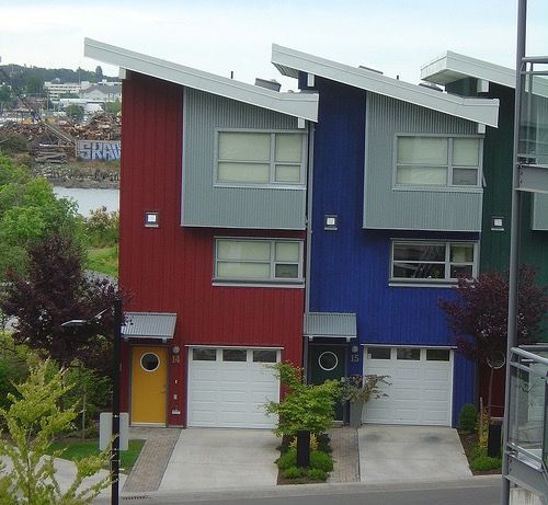 Two townhomes stand side by side. The left house has red metal siding and a slanted metal roof. The right house has blue metal siding and a slanted roof.