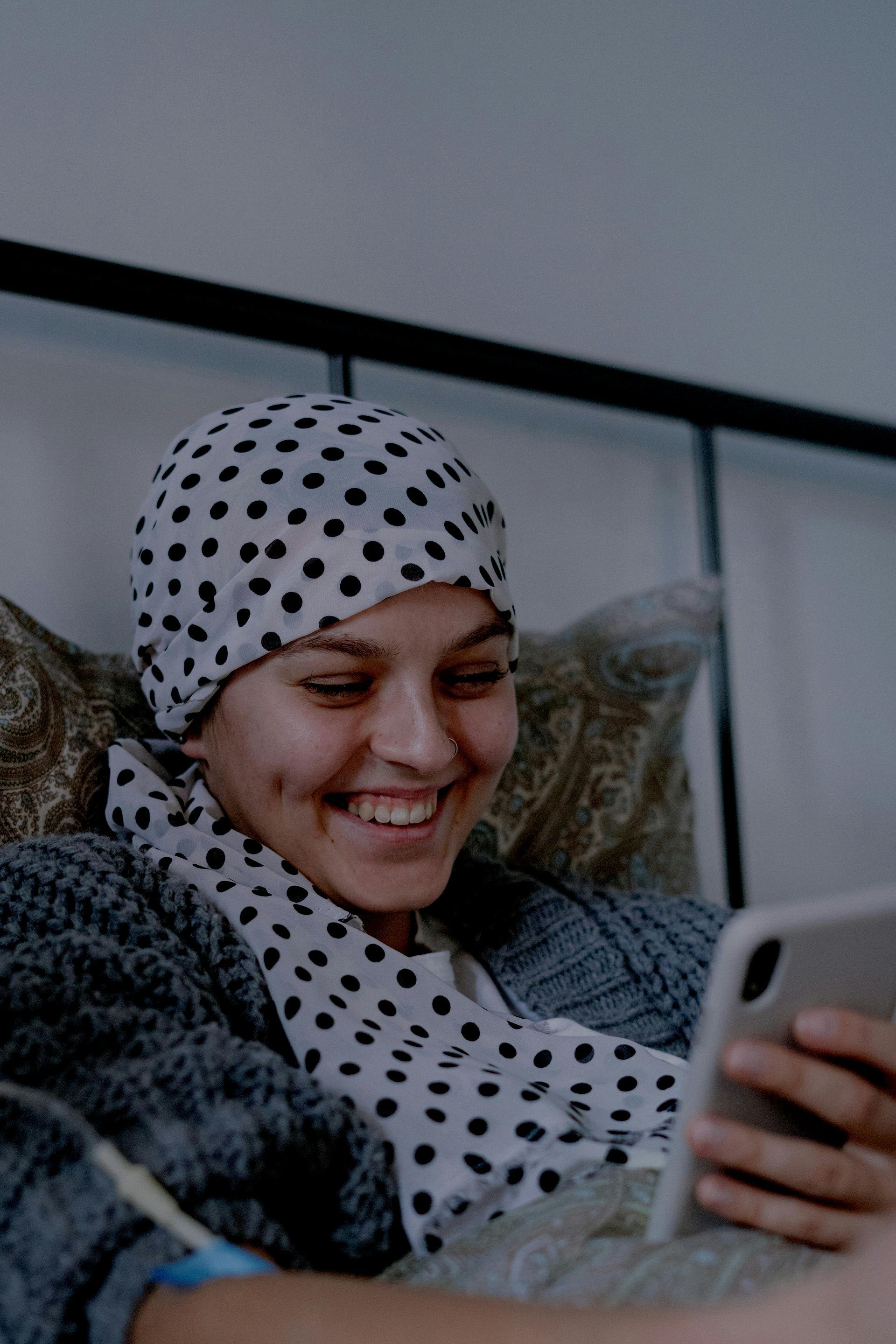 A woman wearing a polka dot hat is laying in bed looking at her cell phone.