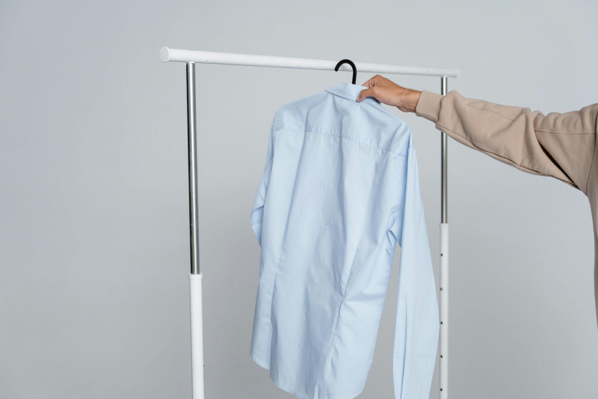 Person's hand holding a blue button-down shirt on a hanger from a clothing rack. Light gray background.