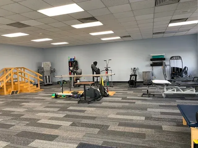 Physical therapy room with equipment: stairs, treadmill, exercise machines, mats. Gray and blue color scheme.