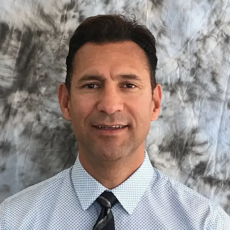 Man wearing a patterned shirt and tie, smiling, against a mottled gray backdrop.