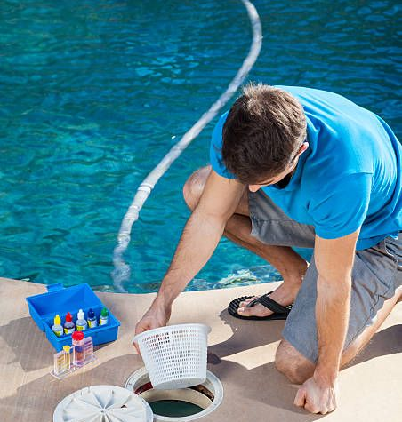 Man crouching beside a pool, checking a basket near the water with cleaning supplies nearby