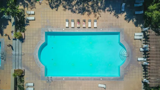 Aerial view of a turquoise swimming pool with lounge chairs on a sunlit deck and trees around it