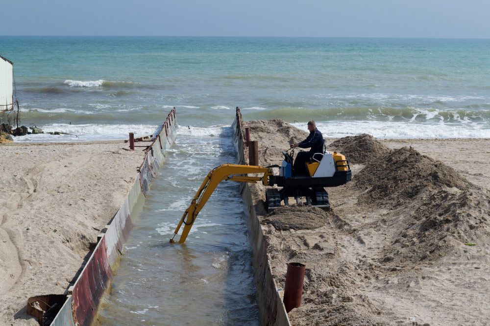 A Man Is Driving A Yellow Excavator On A Beach Near The Ocean — JJ's Excavator & Tipper Hire in Kingaroy, QLD