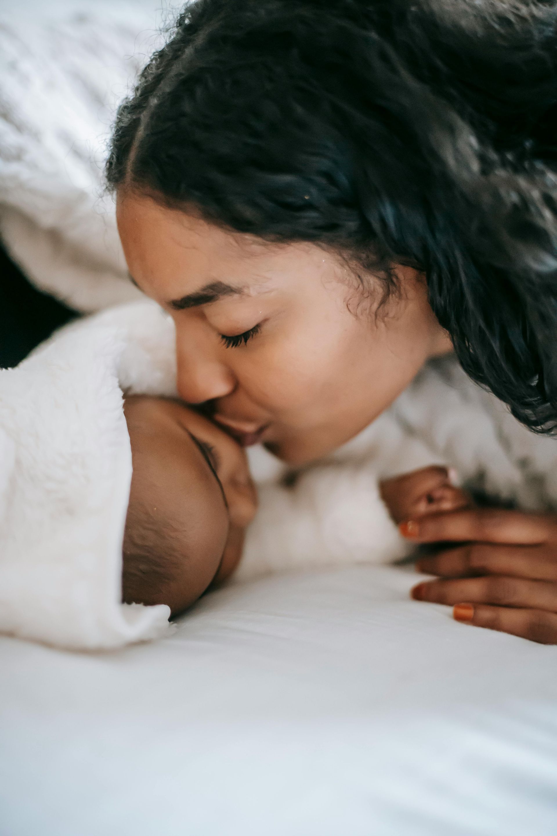 Woman kissing a baby wrapped in a white blanket on a white bed.