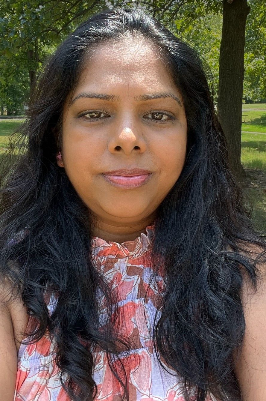 Woman with dark, wavy hair, smiling slightly, in a park, wearing a patterned top.
