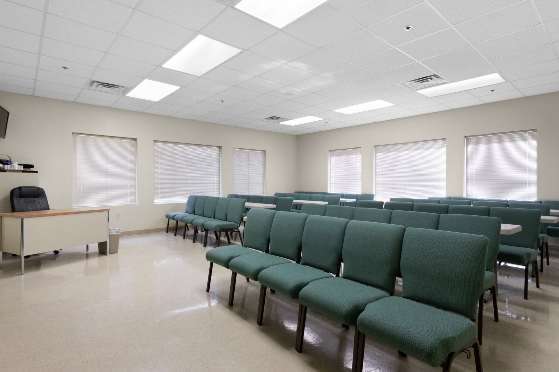 Empty room with rows of green padded chairs, several windows, and a desk.