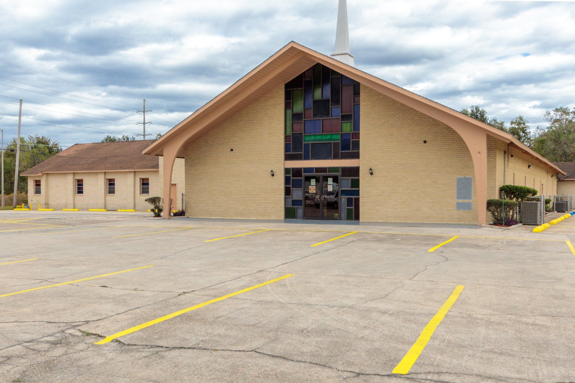 Church building with beige facade, stained glass window, and parking lot under a cloudy sky.