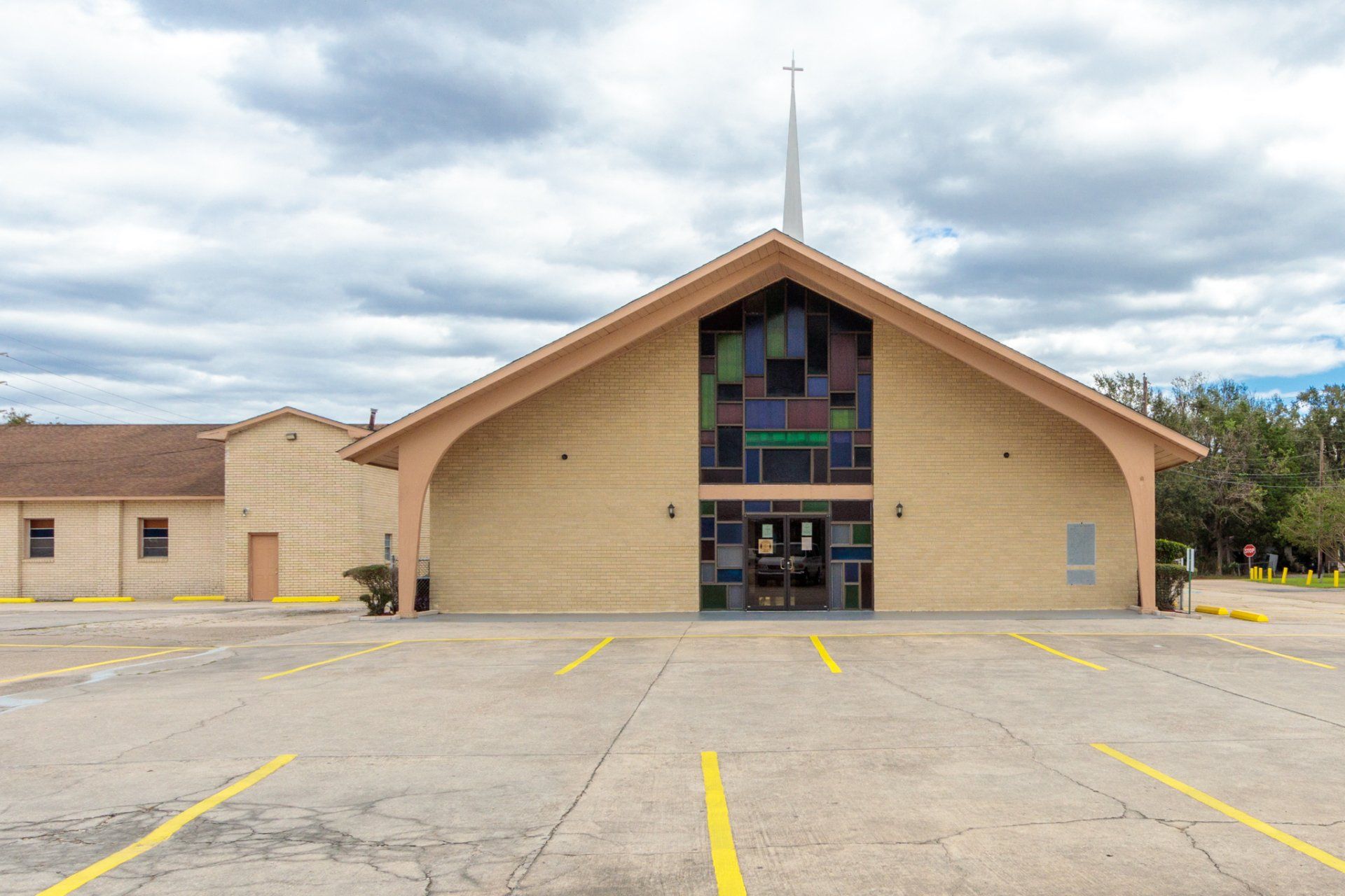 Church building with beige siding, a steeple, and a stained-glass window over the entrance; parking lot in front.