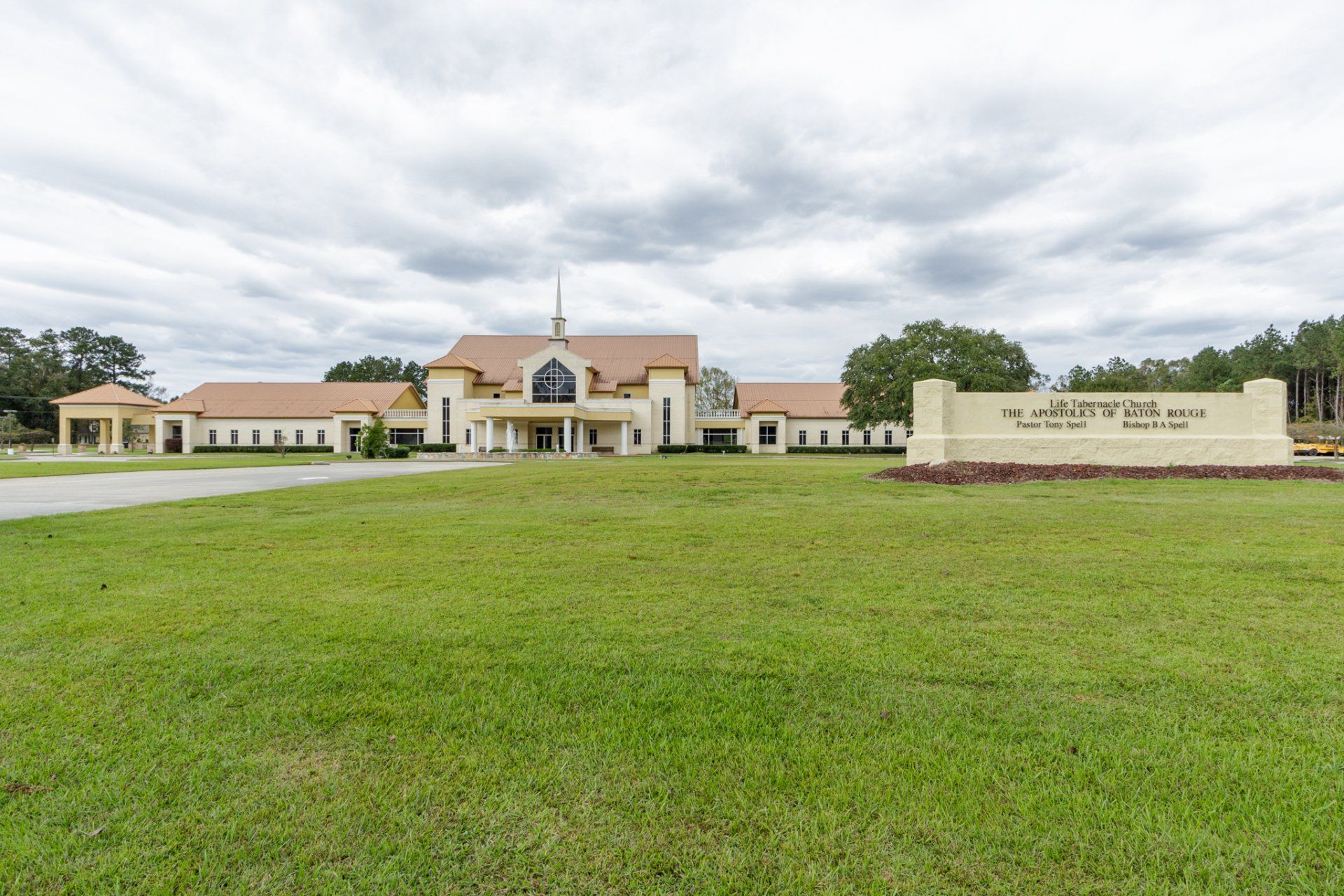 Church building with tan roof and cream-colored facade, with a sign in the foreground. Green lawn and overcast sky.