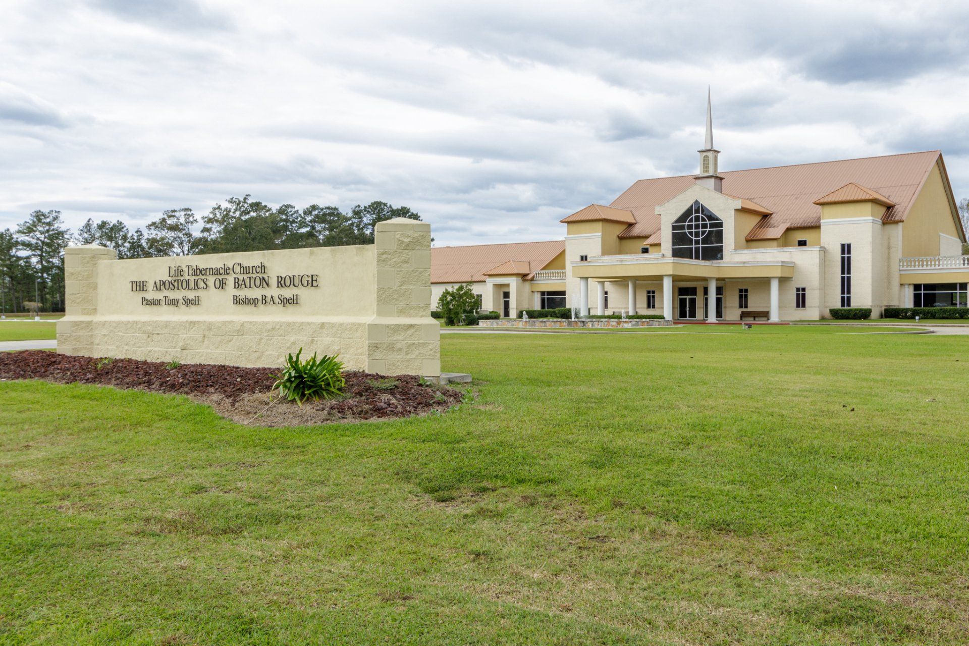 Church building with sign on a grassy lawn under a cloudy sky. The sign reads: 