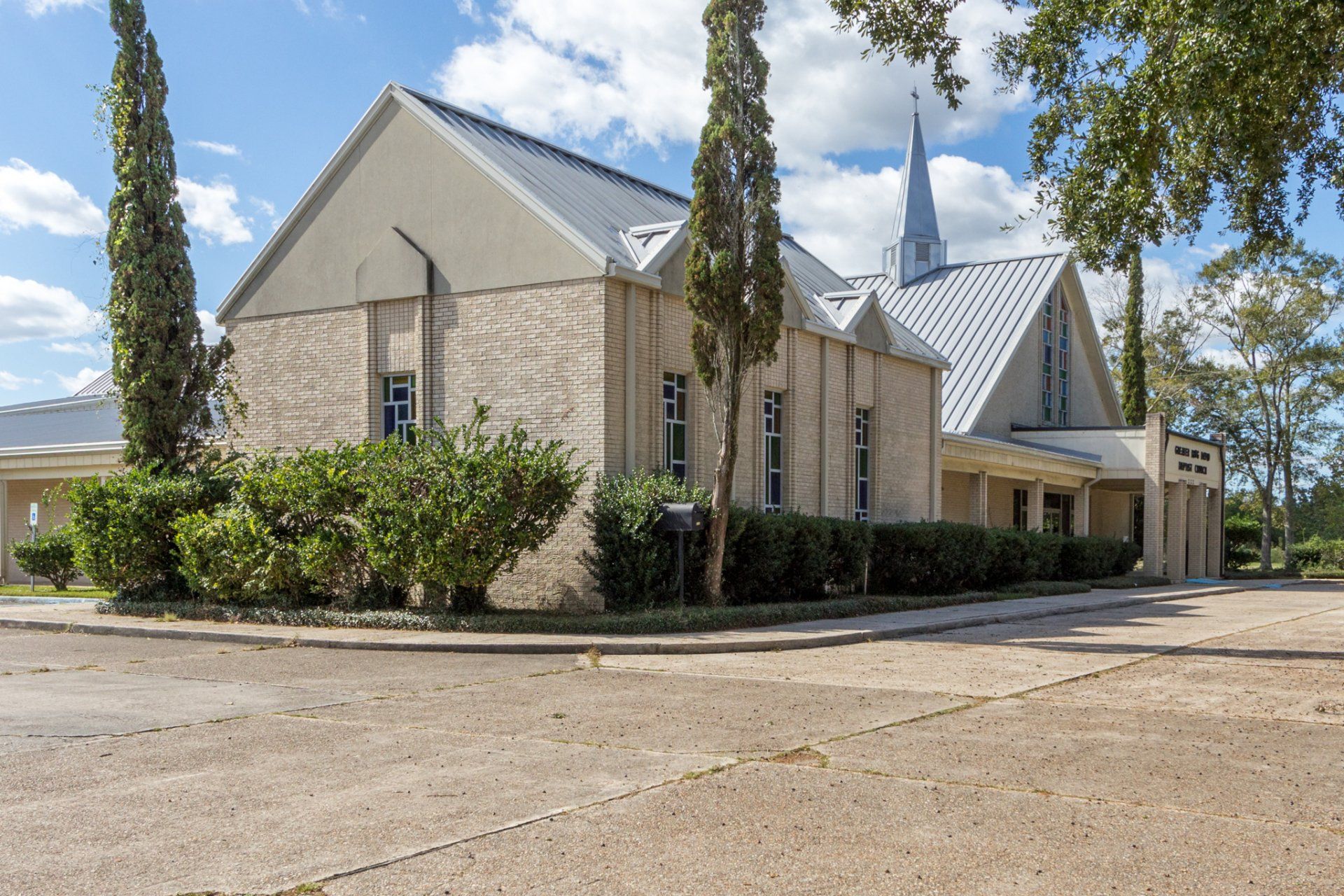 Light stone building with a tall steeple, light blue sky, green trees, and a paved area.