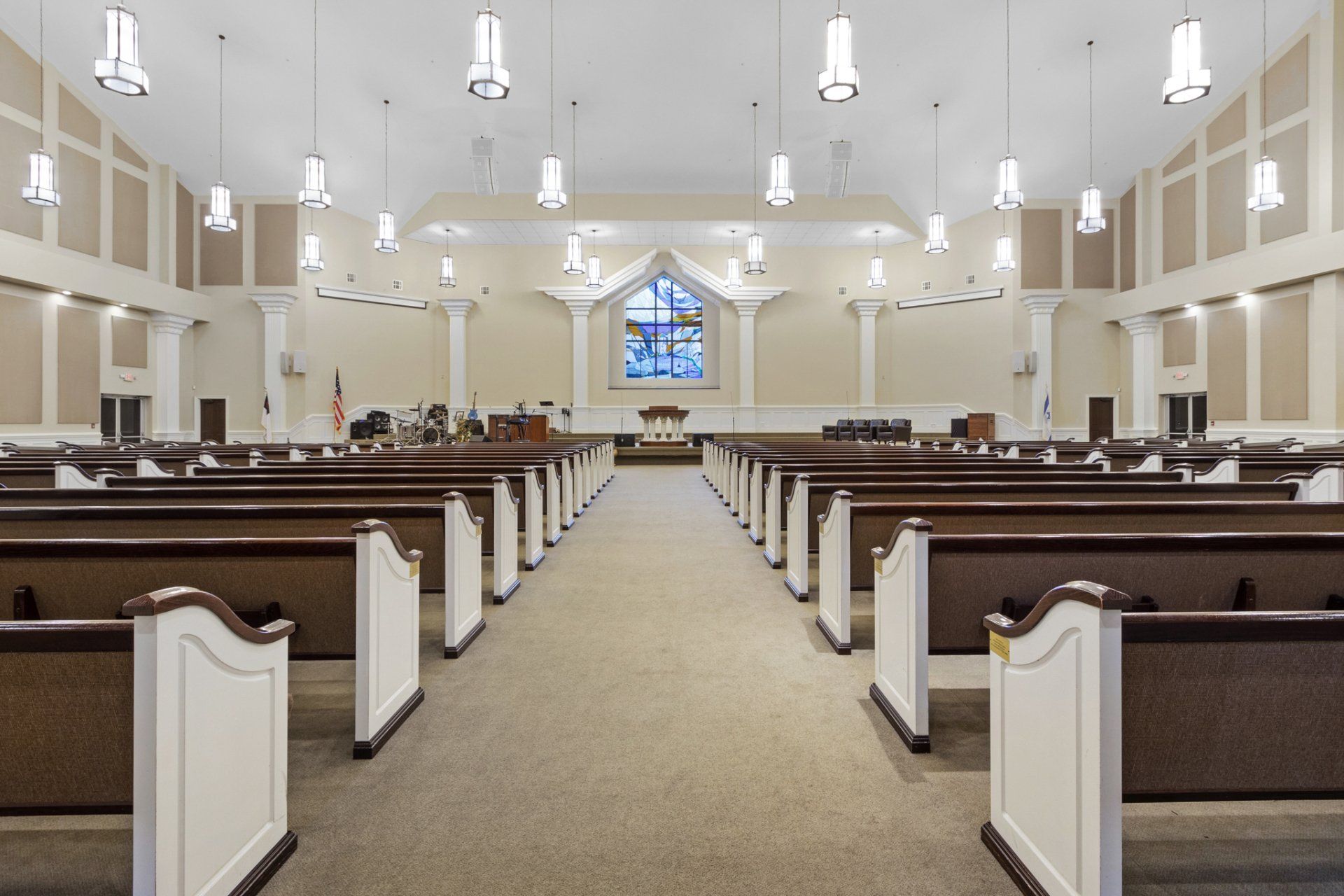 Interior view of a church with rows of pews, a stage, and stained-glass window in the background.