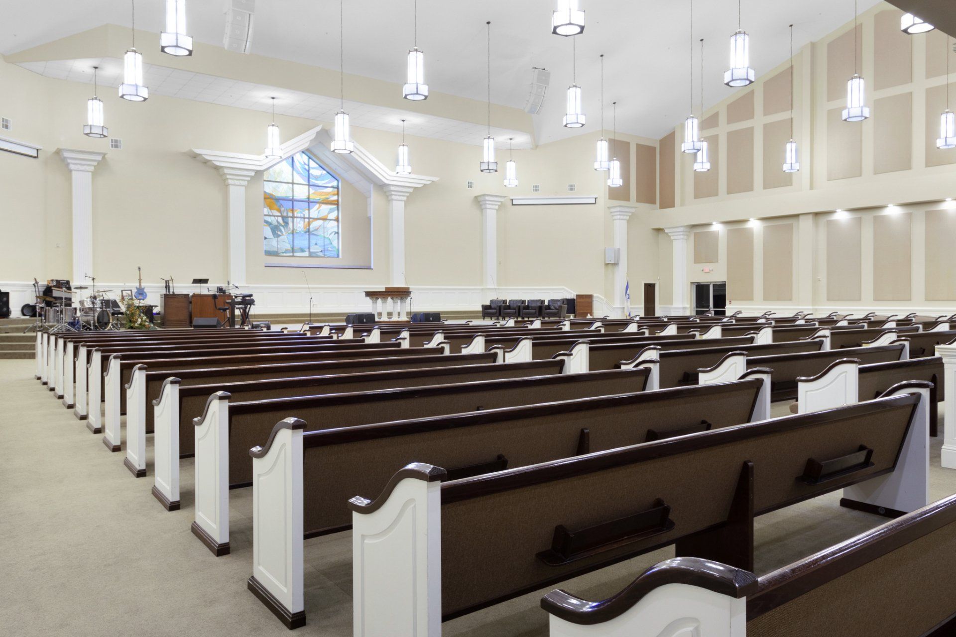 Interior of an empty church with rows of wooden pews. A stained-glass window and stage are at the front.