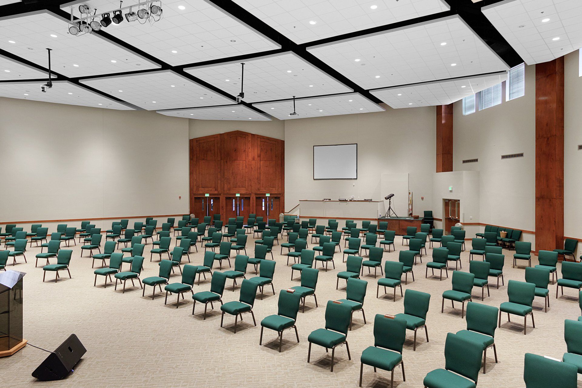 Empty auditorium with rows of green chairs, stage, and sound equipment. Beige carpet.