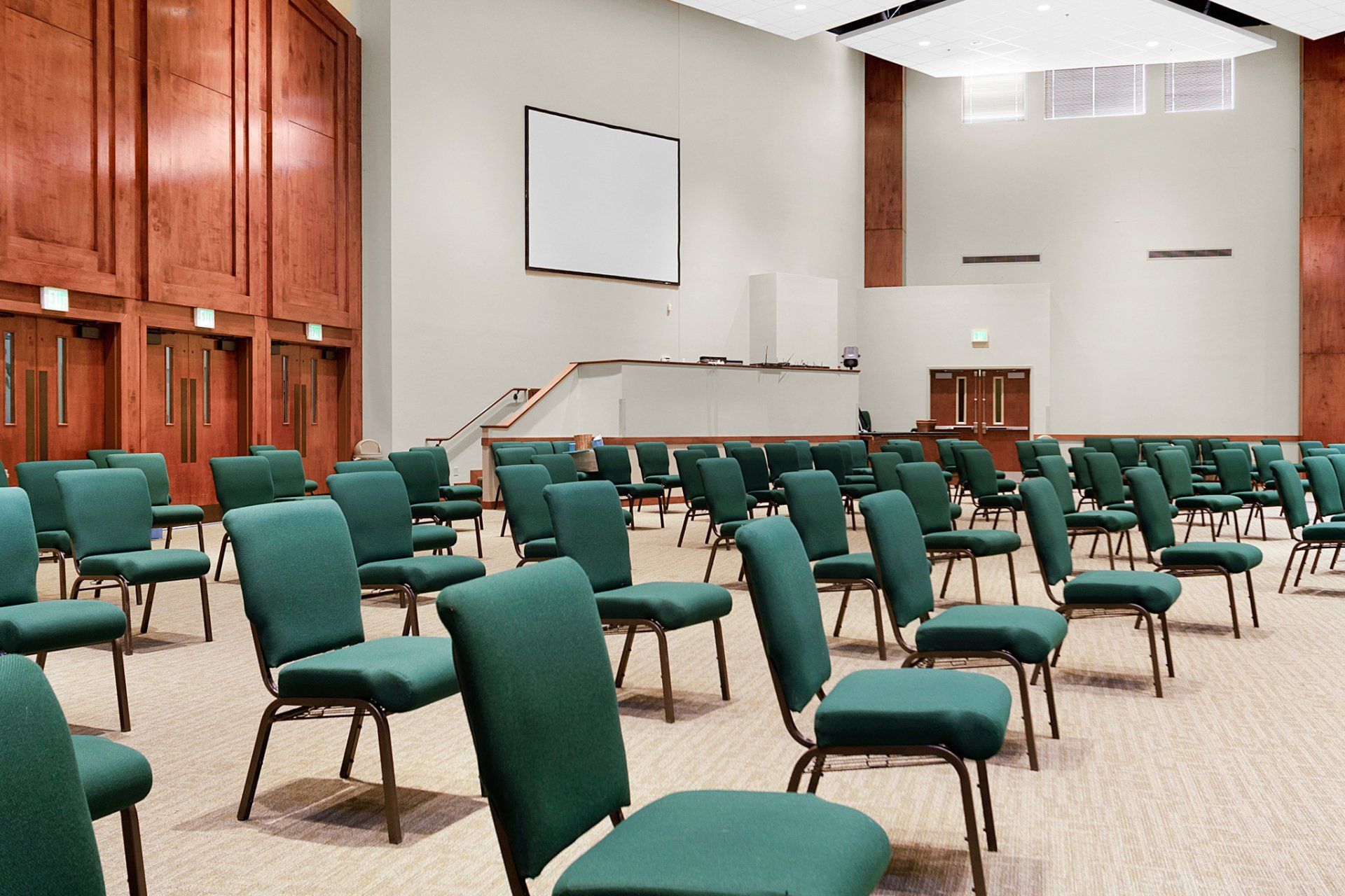 Empty auditorium with rows of green chairs facing a white screen and a raised platform.