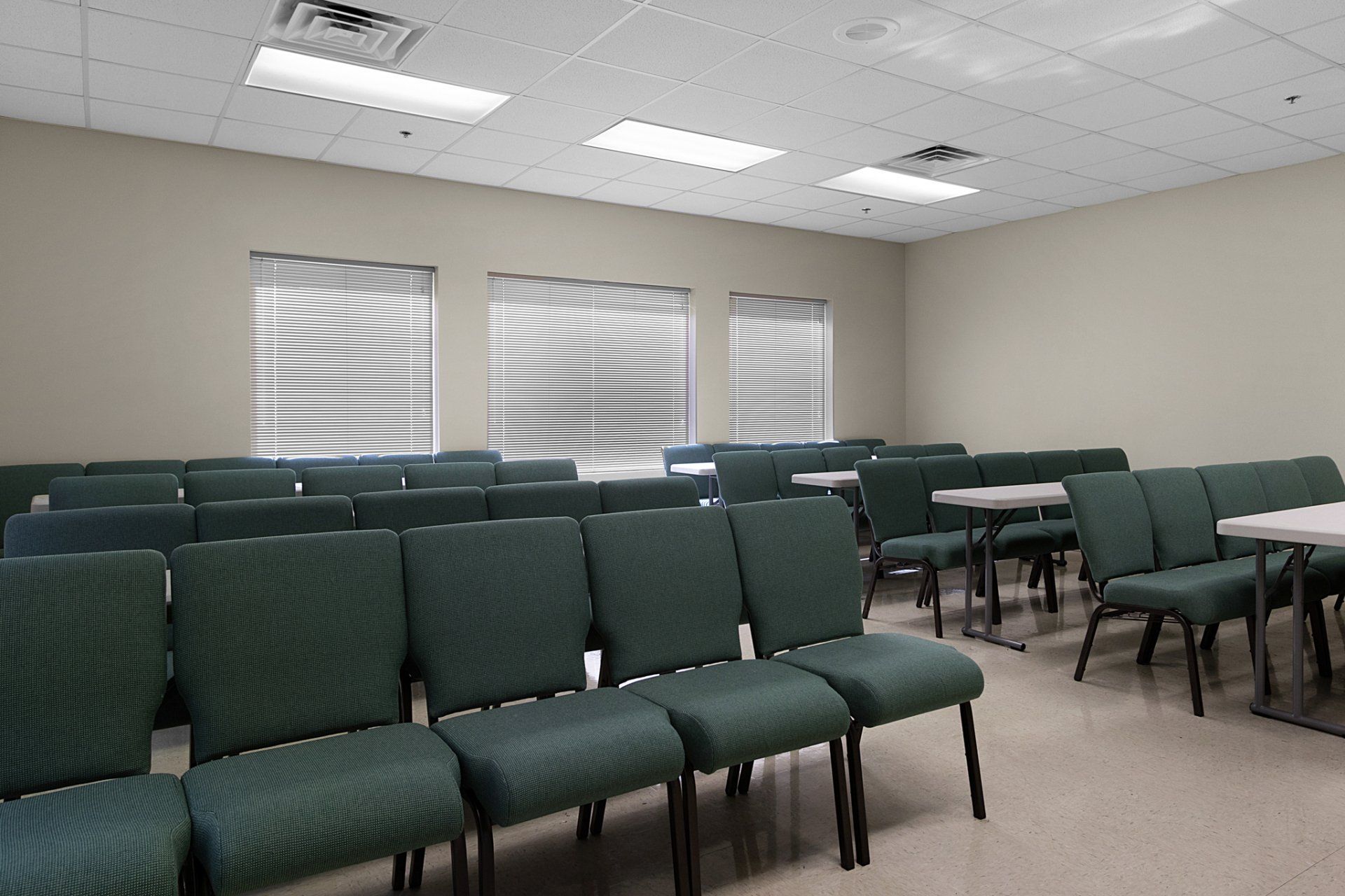 Empty meeting room with rows of green chairs, tables, and window blinds.