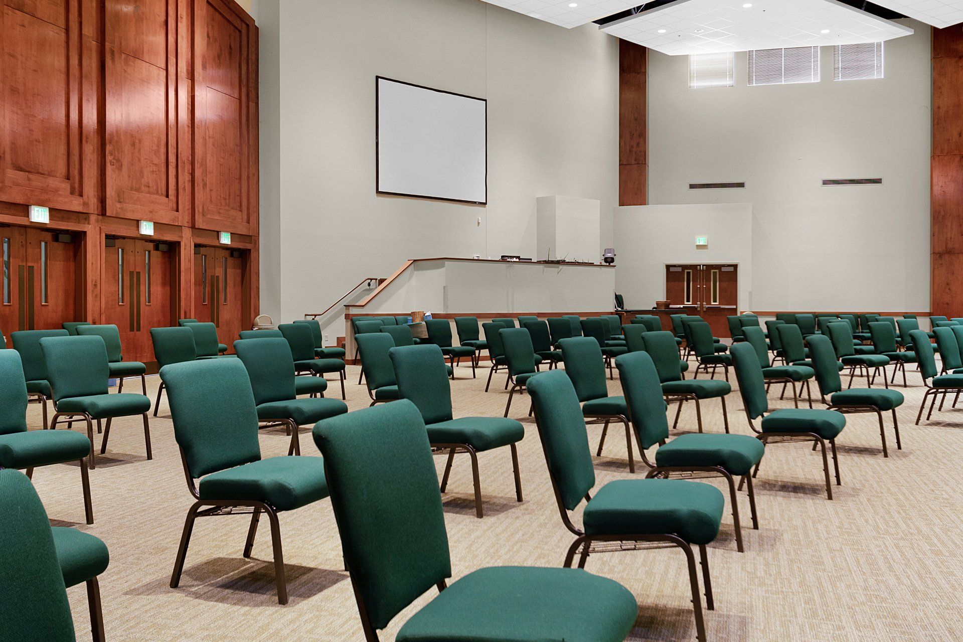 Empty auditorium with rows of green chairs facing a white screen. Wooden walls and beige carpet.