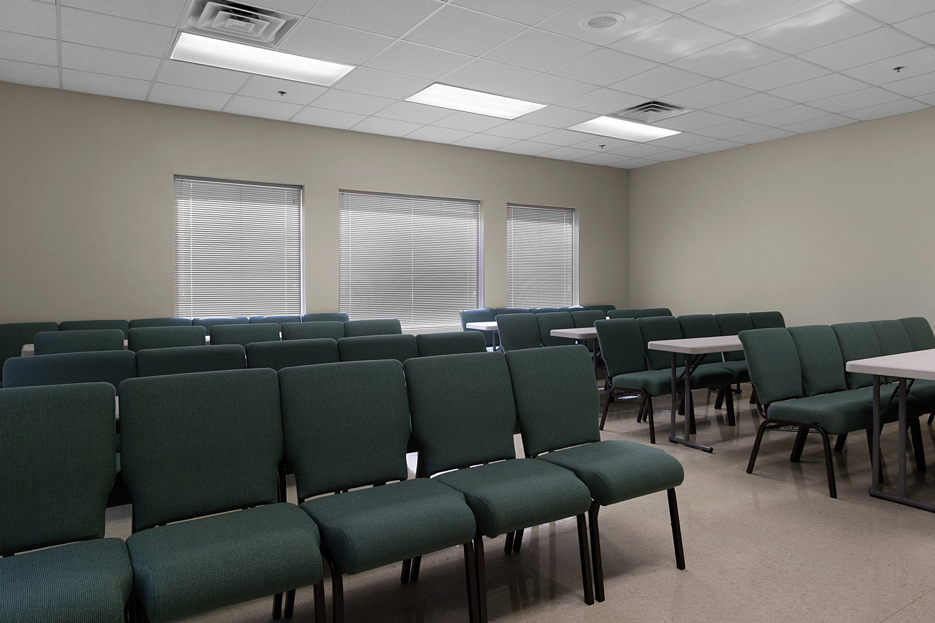 Empty room with rows of green chairs, tables, and blinds on windows.