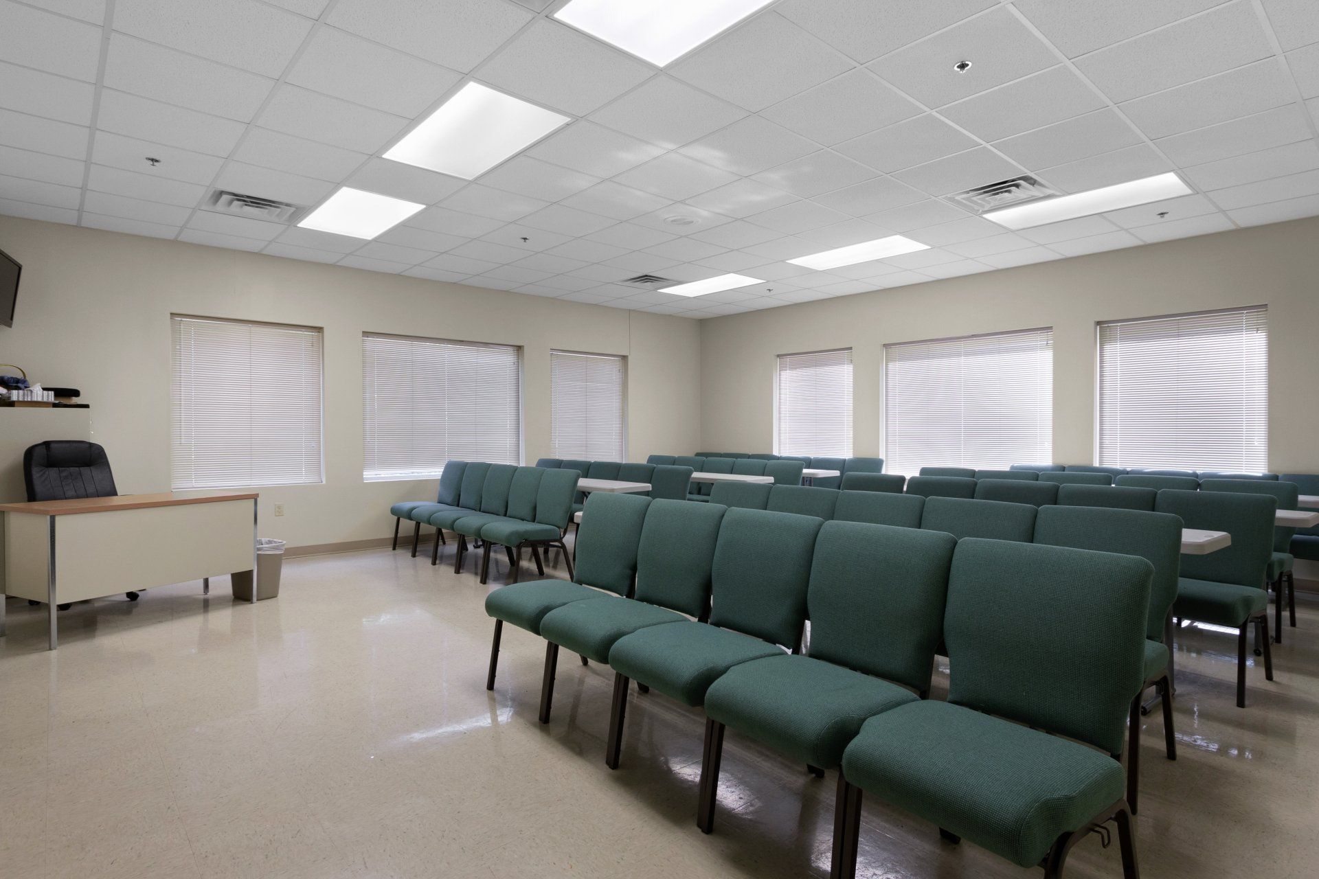 Empty room with rows of green chairs, windows with blinds, and fluorescent lights.