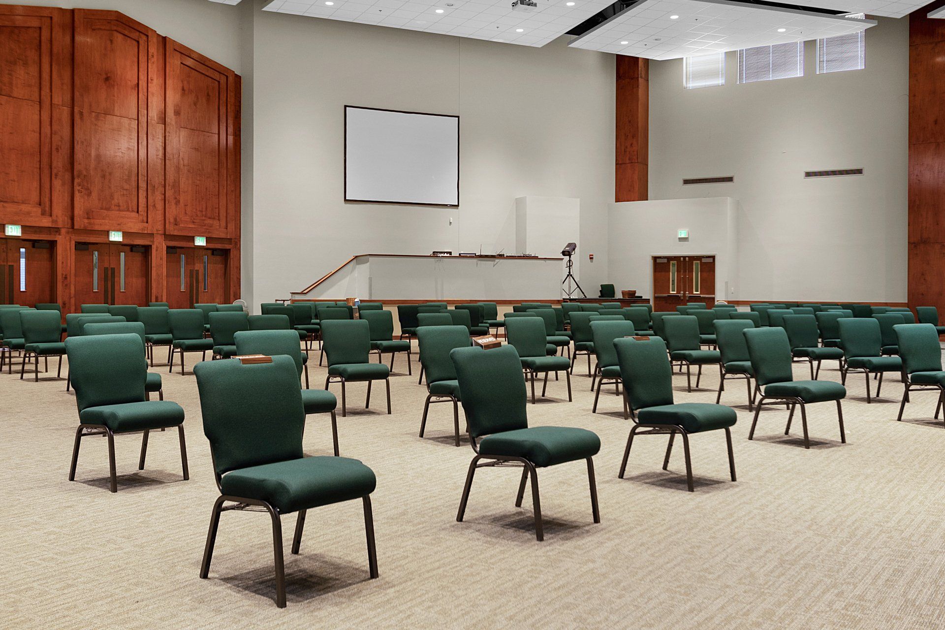 An empty auditorium with rows of green chairs, a white screen, and wooden paneling.