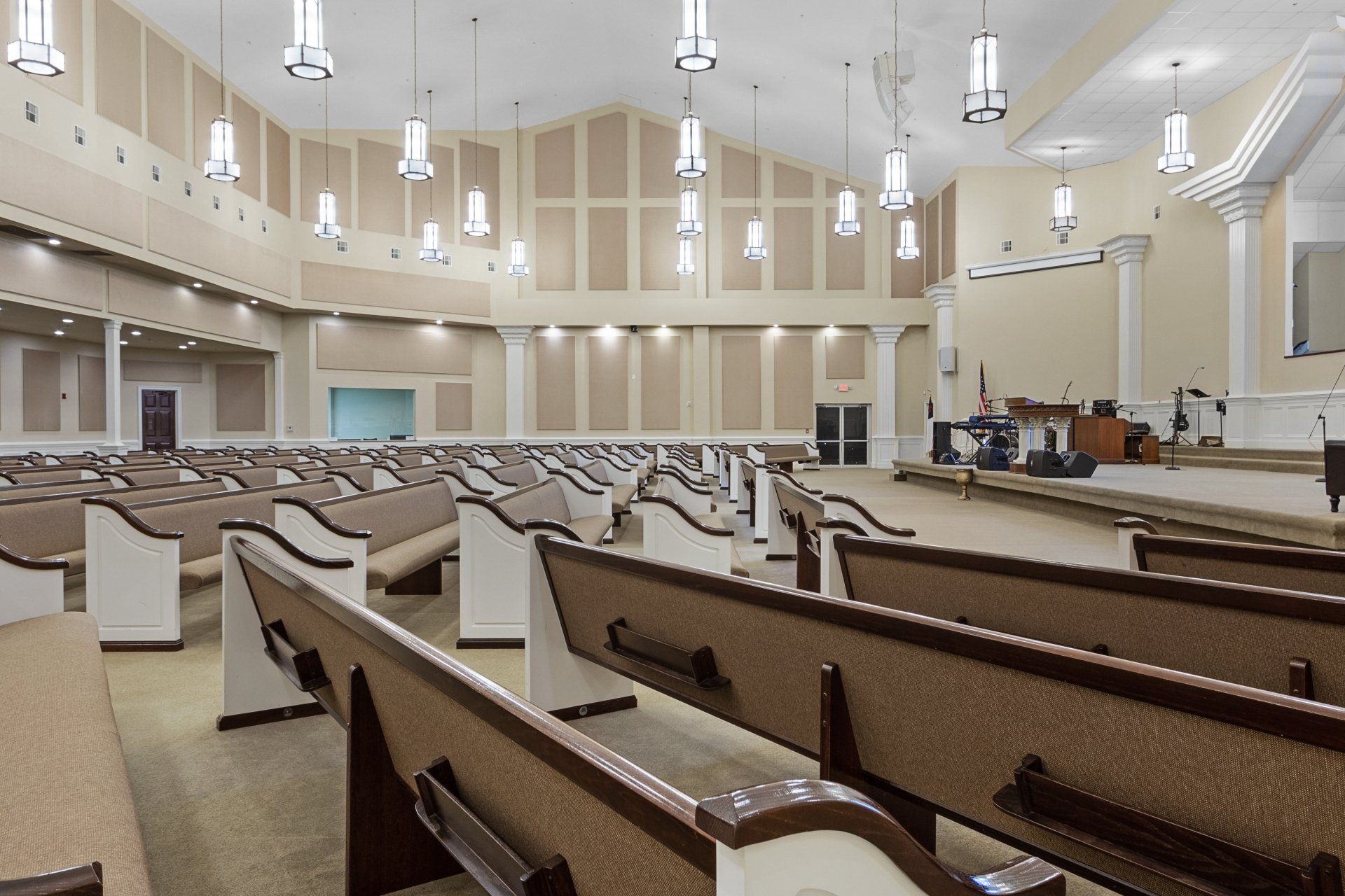 Church interior with rows of pews facing a stage and columns. Hanging lights and acoustic panels on the walls.