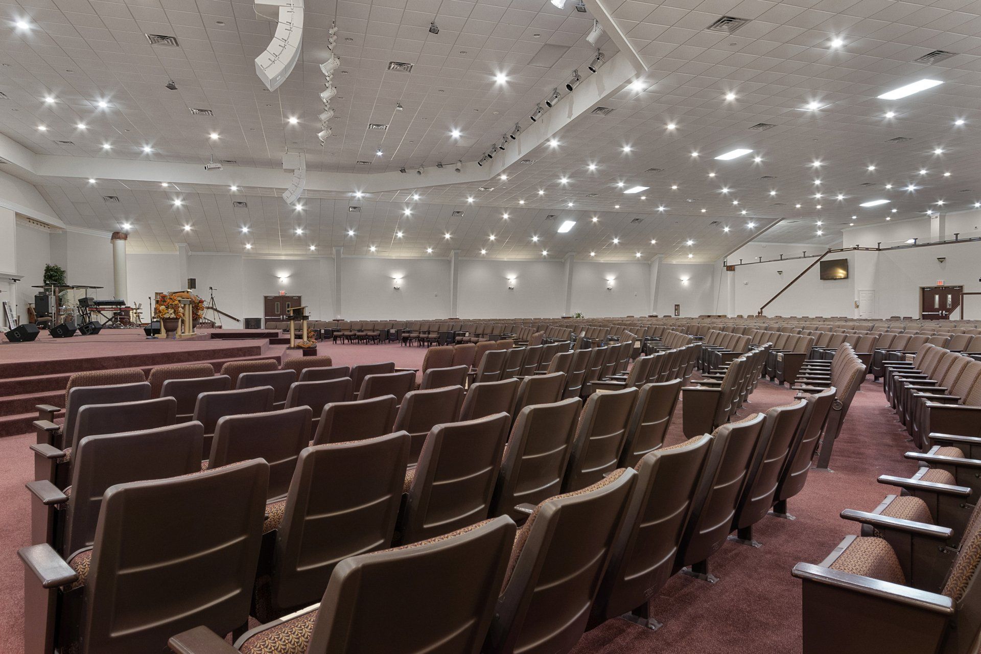 Empty church auditorium with rows of brown seating facing a stage. Bright lights on ceiling.
