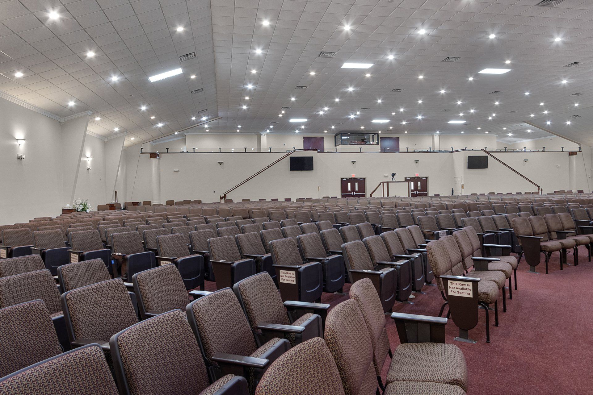 Auditorium with rows of cushioned seats, stage in the back, white walls, and bright ceiling lights.