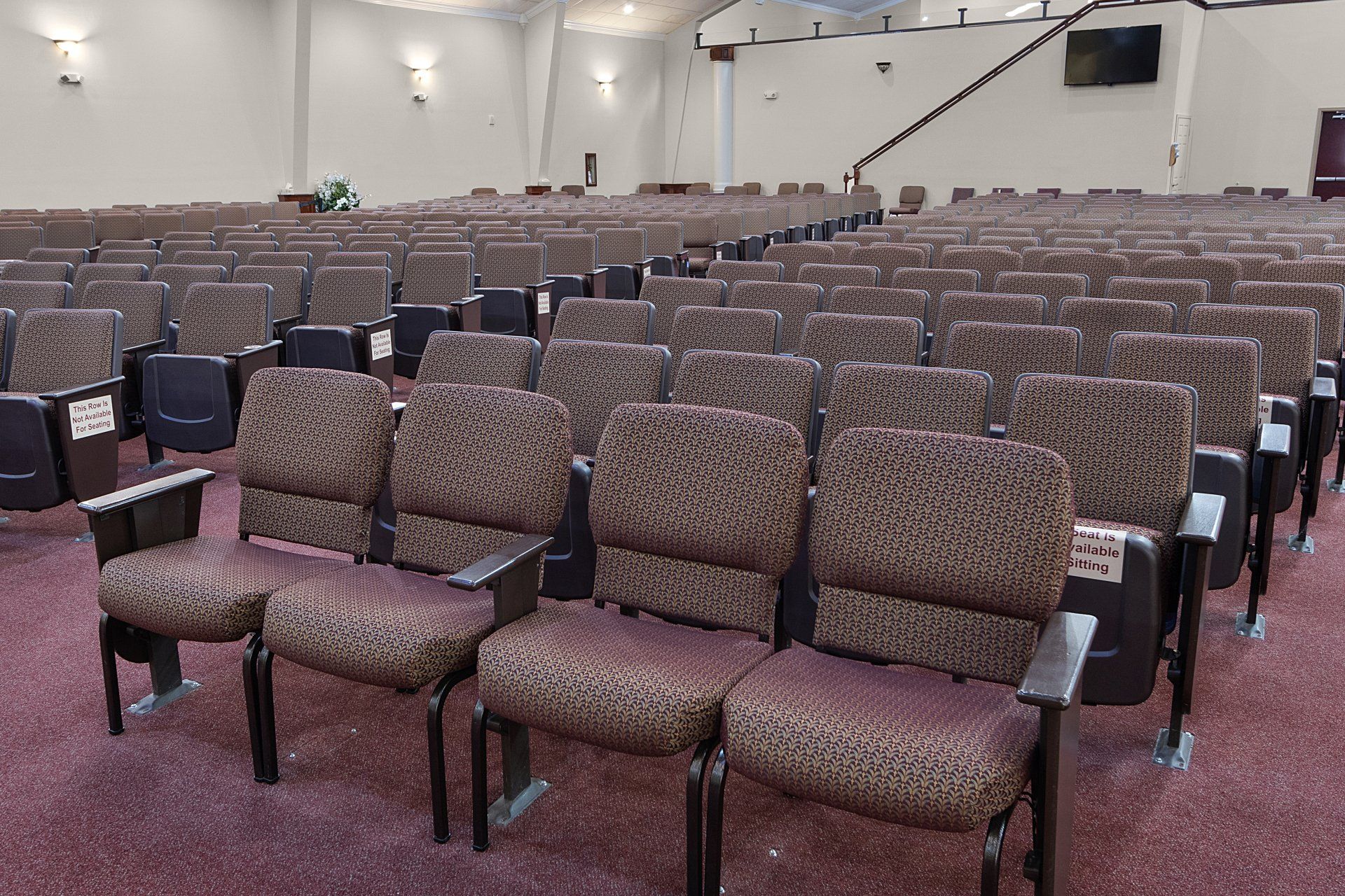 Rows of empty brown patterned upholstered theater seats in an auditorium. Red carpet, cream walls, overhead lights.