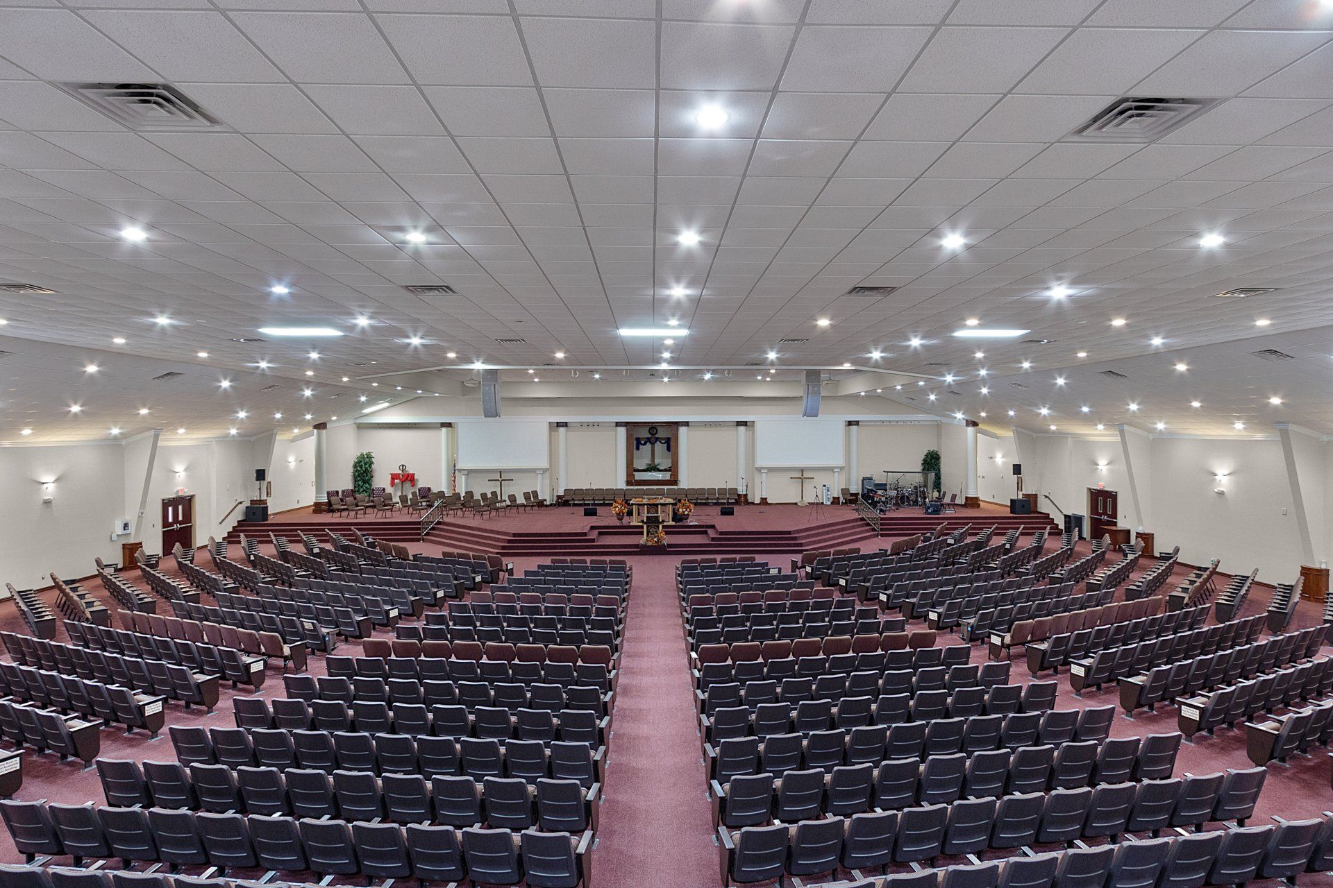 Large auditorium with rows of seats facing a raised stage; burgundy carpet; white ceiling with lights.