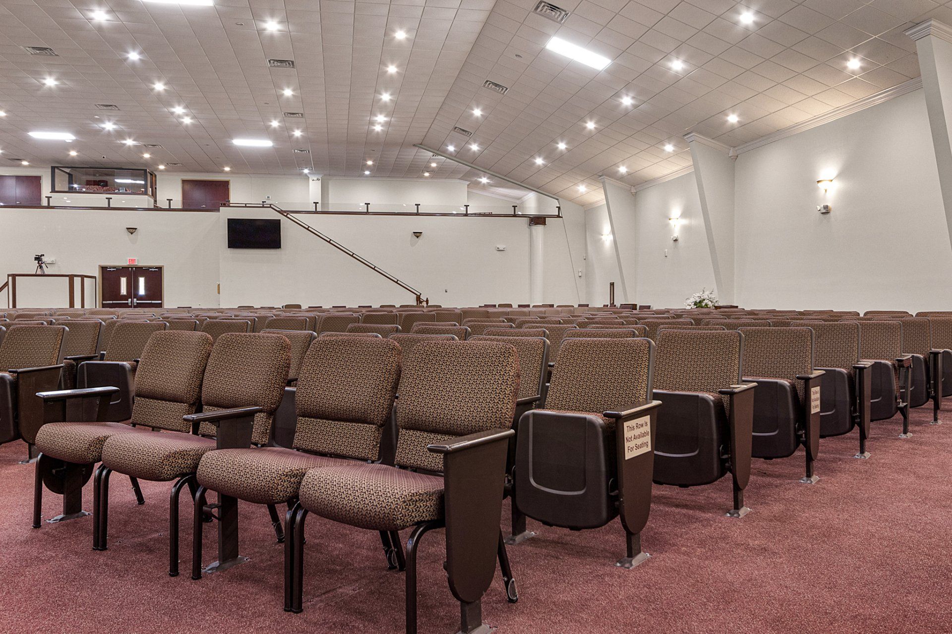 Auditorium with rows of cushioned seats on red carpet, white walls, stage in the distance.