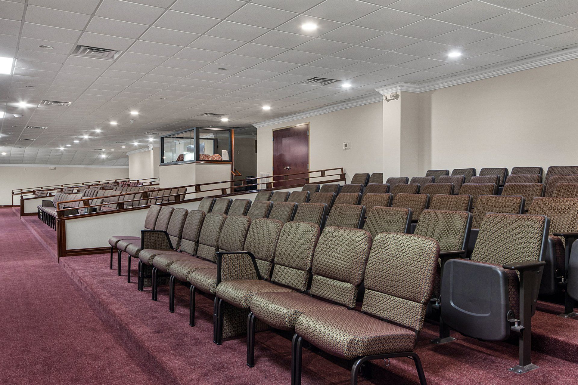 Rows of empty cushioned chairs in an auditorium with burgundy carpet and a neutral ceiling.