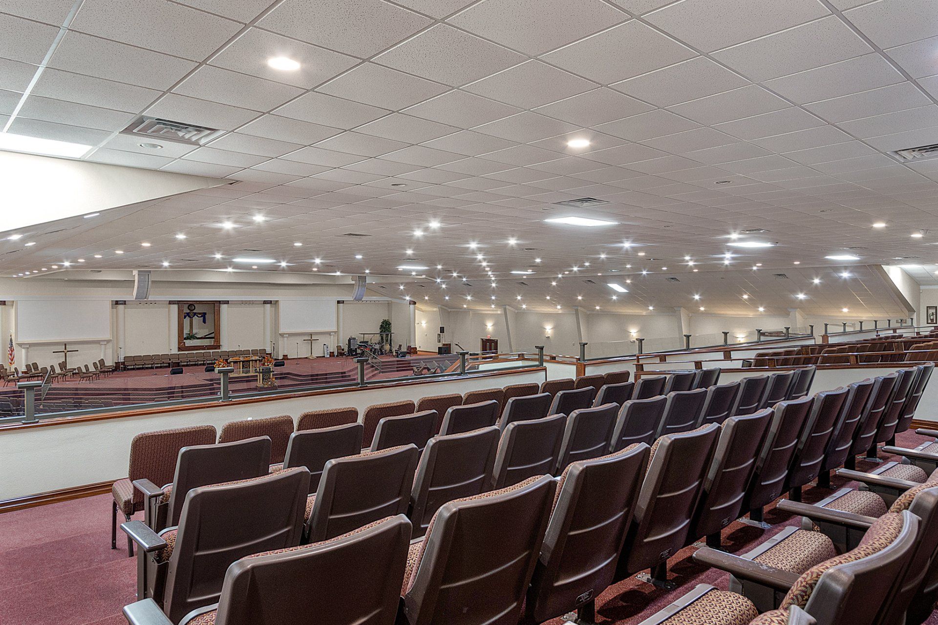 Auditorium with rows of brown seats, stage at front, overhead lights, and a patterned carpet.