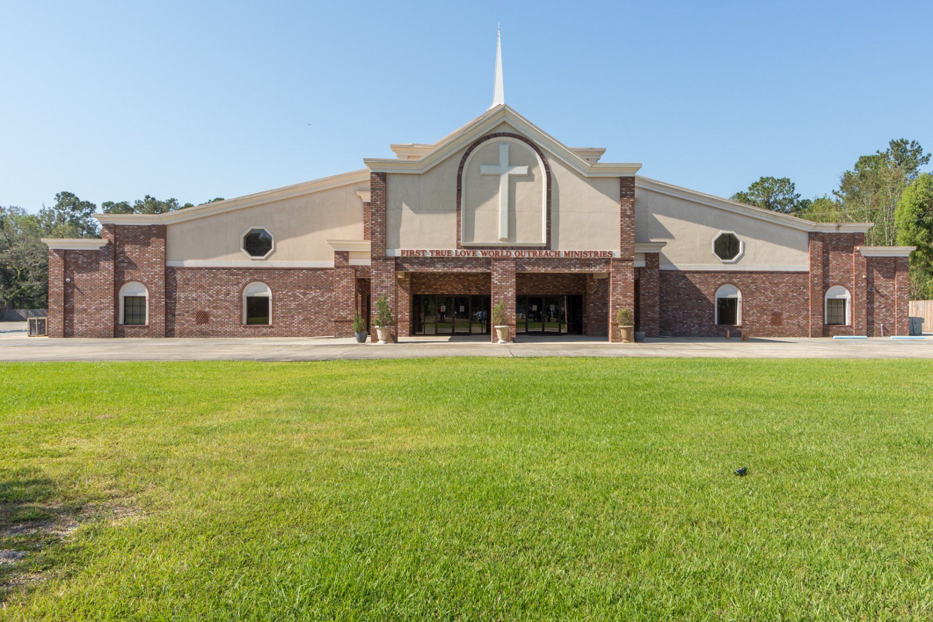Church with light beige facade, brick sides, and cross, set in front of green grass under a clear sky.