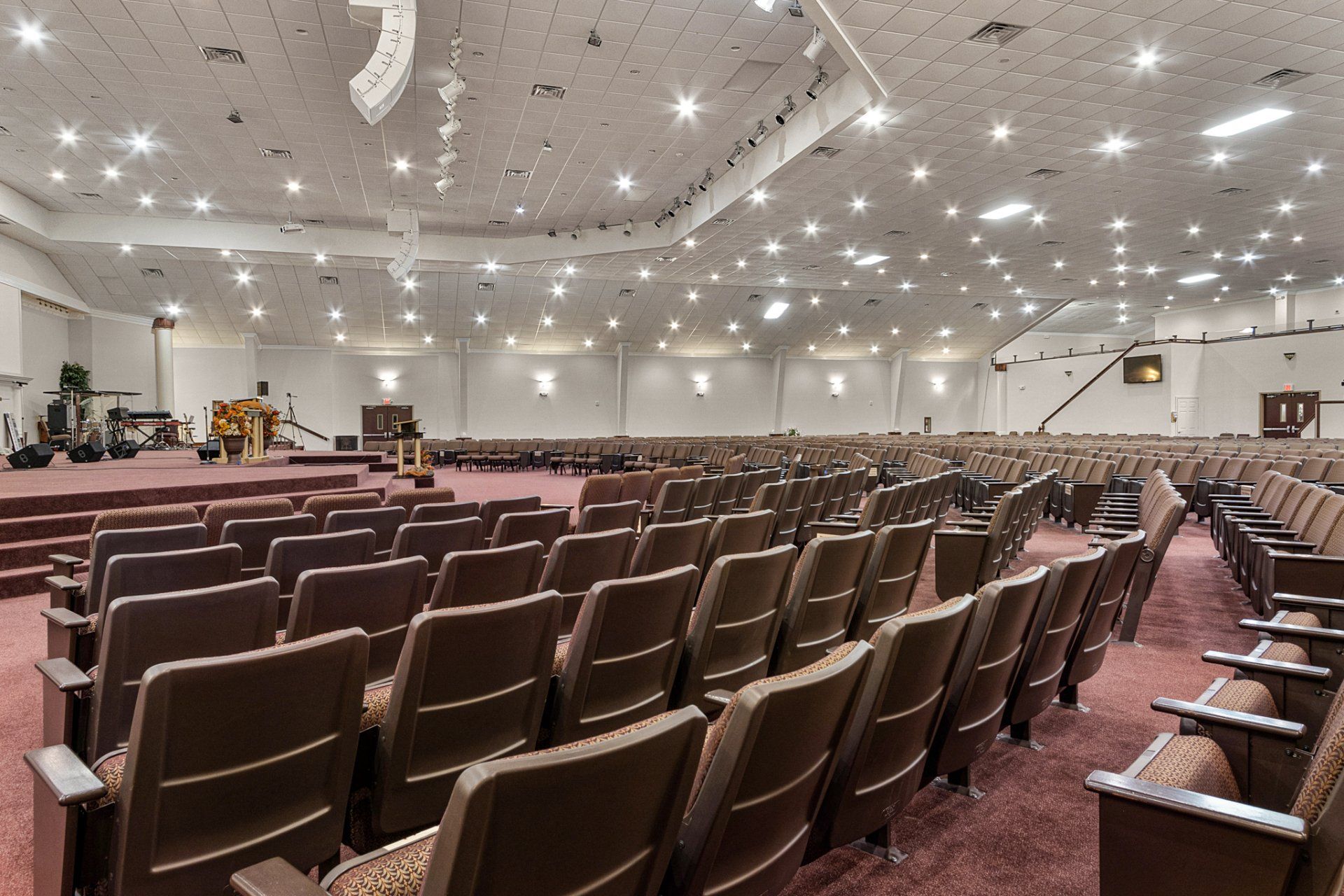 Rows of brown seats fill a large, well-lit auditorium; stage with podium and musical instruments.