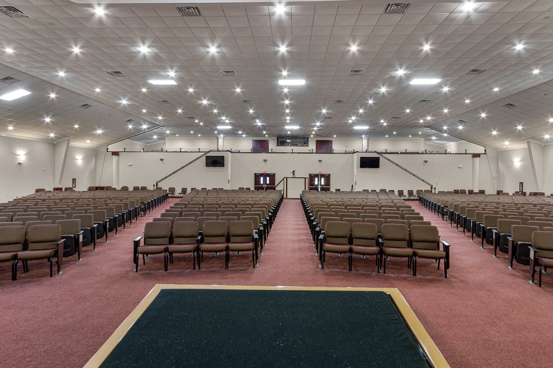 Rows of empty brown chairs in a large, bright auditorium with a stage at the front.