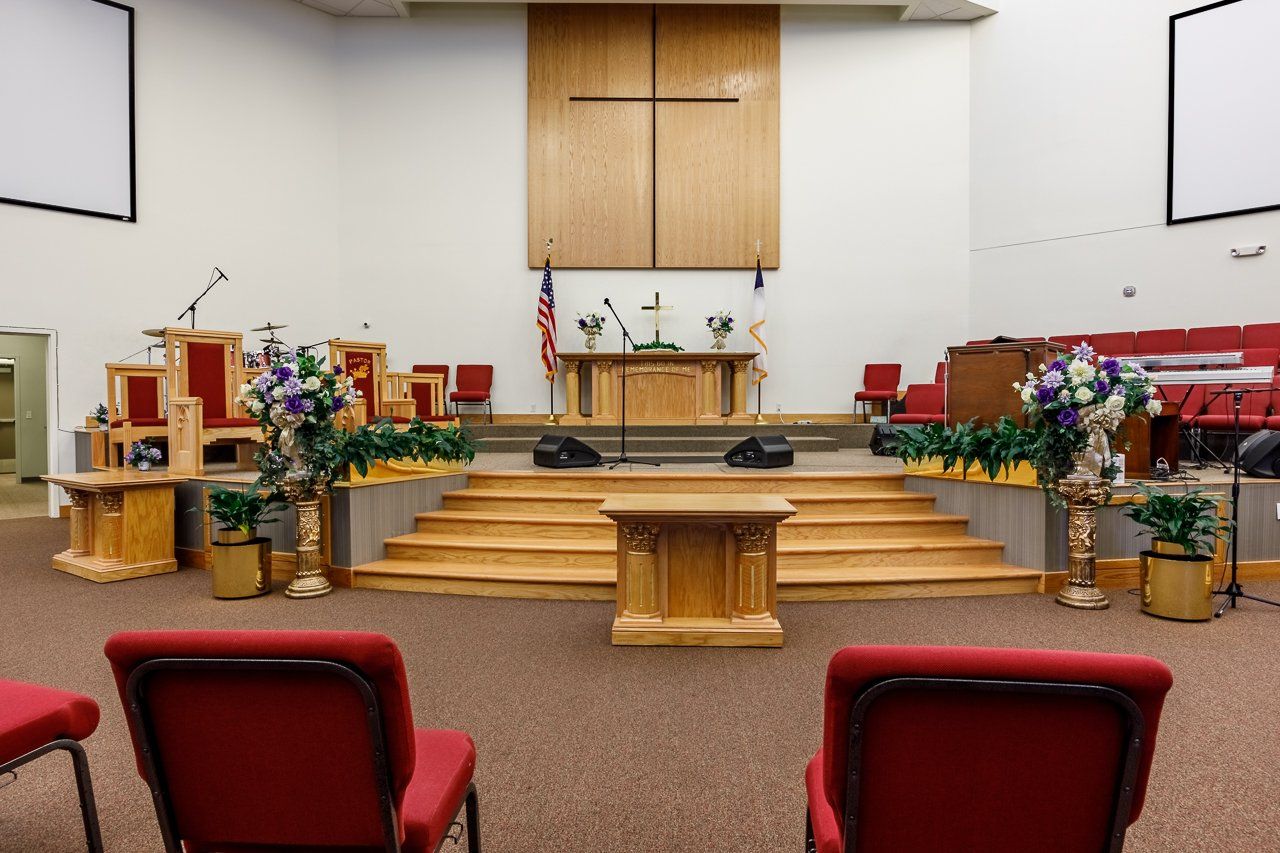 Empty church sanctuary with wooden altar and cross, red chairs, and floral decorations.