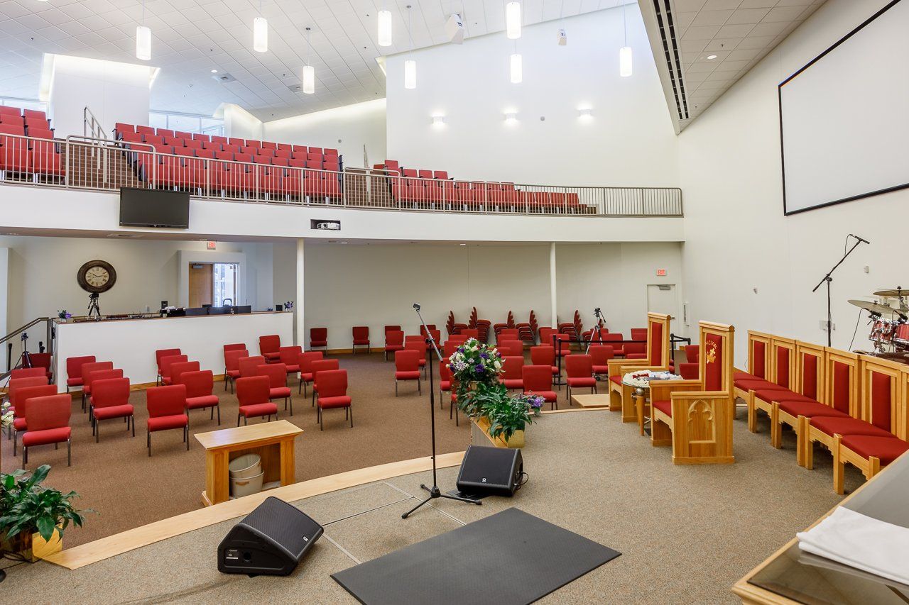 Interior of a church sanctuary with red chairs on floor and balcony, a stage with equipment, and white walls.