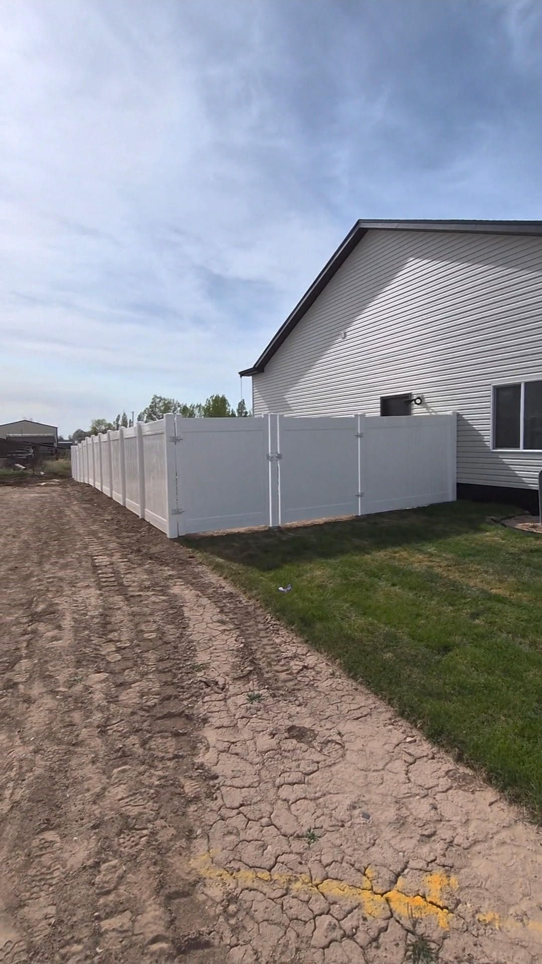 White fence beside a house along a muddy dirt path under a partly cloudy sky