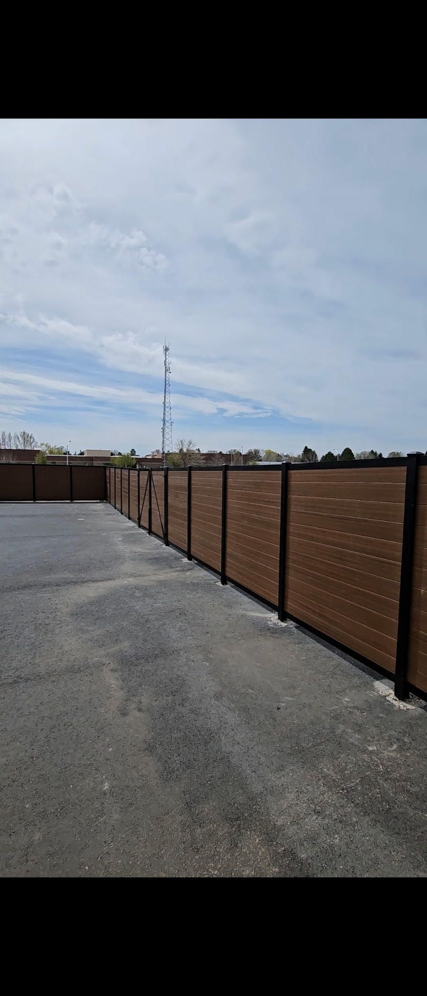 Gravel lot beside a long brown fence under a cloudy sky with a distant cell tower
