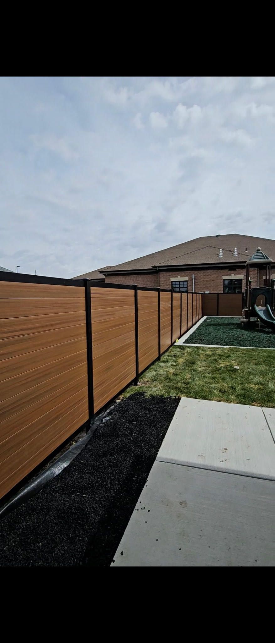 Backyard walkway beside a wooden fence, black mulch beds, and a house under a cloudy sky