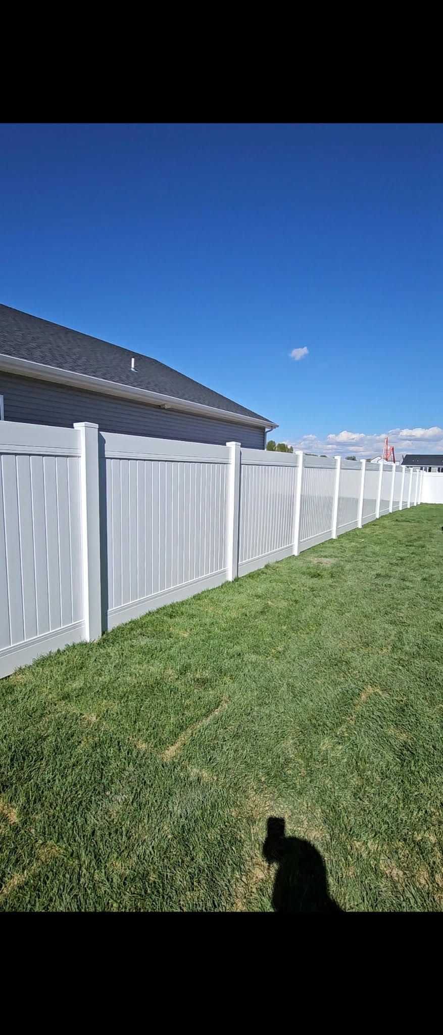 White fence beside a green lawn under a clear blue sky, with a house roof at left and a small cloud overhead