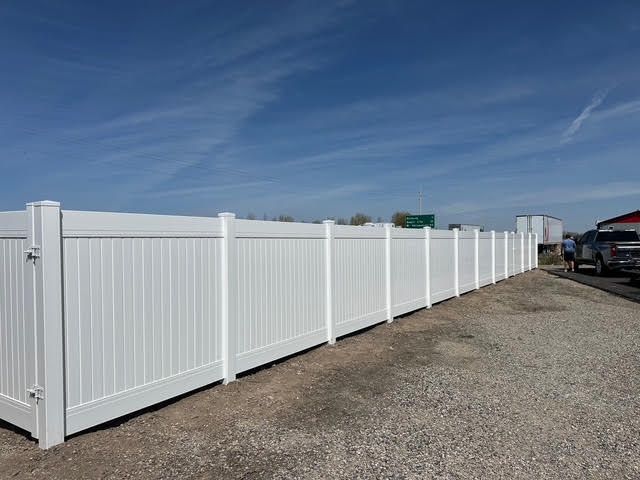 White vinyl fence along a gravel roadside under a blue sky, with parked vehicles in the distance